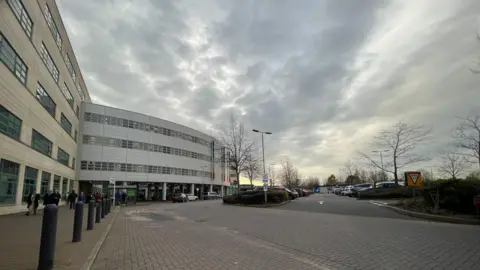 A view of a hospital building with a car park to the right side. It is a cloudy day.