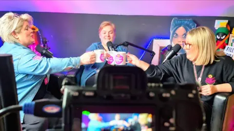 Support 2gether Three women smiling sitting in a triangle. They are holding mugs that have the pink logo of Support 2gether on them. The three women have blonde hair and have podcast mics in front of them. There is a black camera with the image blurred in the view finder. 