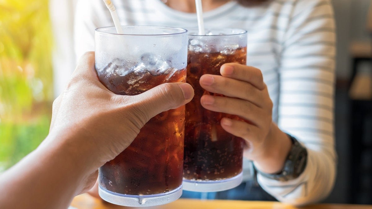 Two people holding and clinking glasses of sofa with straws and ice seen inside