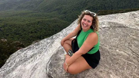 Handout A woman wearing white sunglasses, a green tank top and black skirt squatting on a cliff ledge with thick treetops on the horizon. 