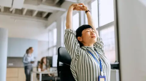 Getty Images A busy woman takes a moment to stretch while working at her desk in a modern office, balancing productivity and well-being