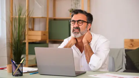 Getty Images A man with a laptop on his lap is hunched over and rubbing his neck. He is in pain and is likely working on the computer.