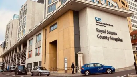 BBC The exterior of the Royal Sussex County Hospital in Brighton, with the name of the hospital written on the side of the building. A pedestrian is walking into the entrance, being passed by a blue taxi. Several other vehicles are parked on the side of the world.