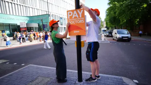 Press Association A man and a woman wearing orange BMA caps attach at Pay Doctors placard to a post on a traffic island outside Bristol Royal Infirmary. The entrance of the hospital, with pickets and media outside, is visible in the background on the left side of the shot, while cars queue on the right hand side. 