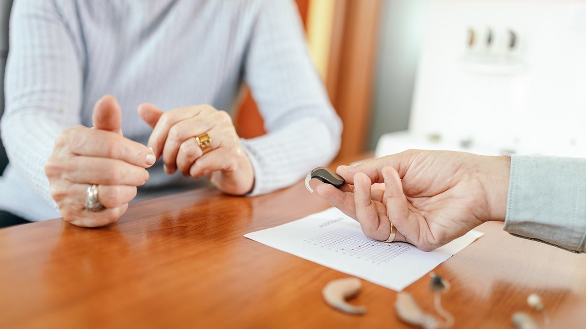 Senior woman choosing hearing aid device in specialized medical clinic. Doctor showing different kinds of devices. Medical test results carton is on the desk in front of her. A study shows that repairing hearing loss can improve senior loneliness