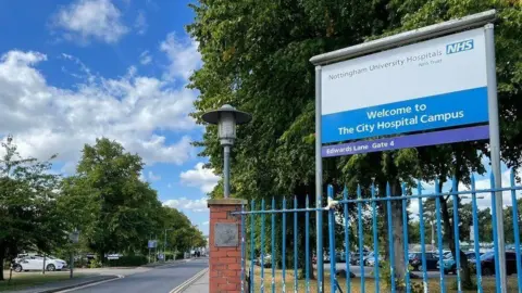 LDRS The entrance to the City Hospital Campus, with a tree-lined road stretching into the distance on the left and blue metal railings and a sign for the hospital to the right