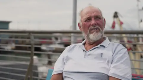 Rob Caunter, 66 years old, is seated and staring off camera to the right of frame. He is sitting down on a bench in the port with fishing boats behind him. He has white hair and a white beard and is wearing a grey and white polo T-shirt.