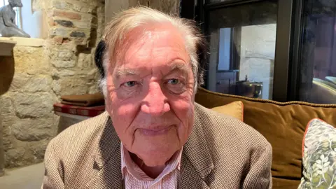George Davies is sitting on a sofa and leaning towards the camera. He is wearing a red and white shirt and beige jacket. He has grey hair and he is smiling.