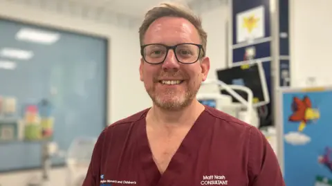 Consultant neonatologist Matt Nash is standing in a hospital area. He is wearing maroon scrubs with his name on his top. He has a beard and is wearing glasses. There is hospital equipment behind him and a brightly coloured picture.