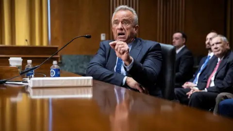 Politico AP Robert F Kennedy, sits at a wide wooden table. He is wearing a smart, tailored, dark suit. On the table is white folder, some plastic bottles and a white disposable drinks cup. He is speaking animatedly and gesturing with his left hand. In the background, other men in similar dark suits listen. 