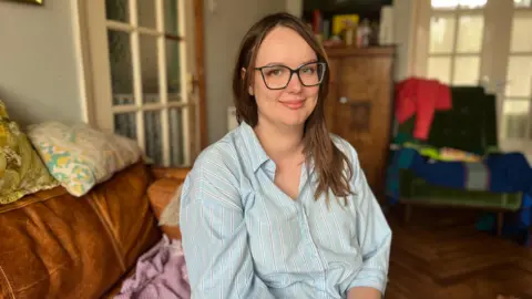 BBC Elissa in a pale blue striped shirt sitting on a leather sofa, wearing glasses with her hair down