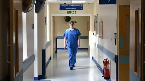 Getty Images A doctor in blue scrubs is walking towards the camera. She is in an NHS hospital corridor with doors on both sides and a blue sign above her saying 'waiting area'.
