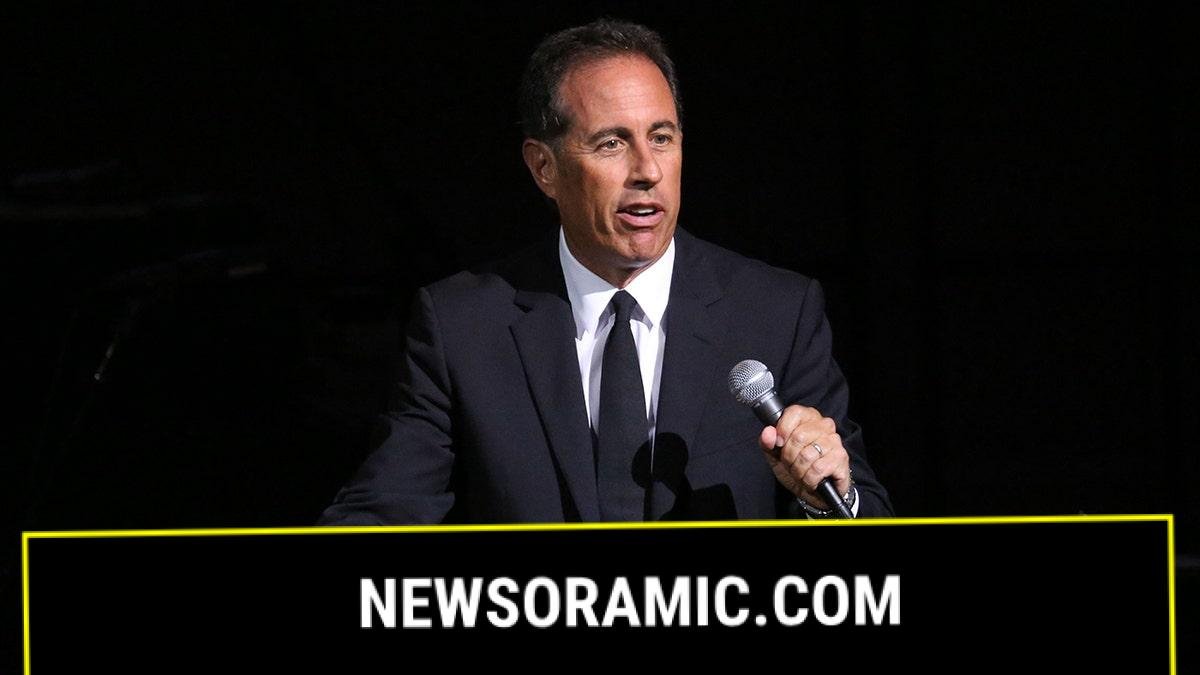 Jerry Seinfeld in a black suit and tie holds a microphone and performs on stage