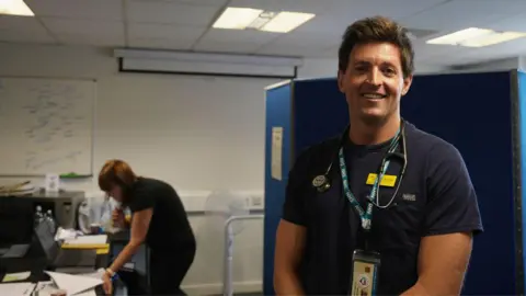 Dr James Gunning, an NHS GP, stands in the temporary fishermen's health clinic in Brixham. He is wearing an NHS T-shirt and has a stethoscope around his neck. Behind him is a receptionist looking at her computer and booking patients into the clinic who turn up for health checks.
