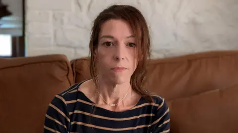 Robby West/BBC Emma Simmonds is sitting down on a brown sofa. She is looking directly at the camera and is wearing a navy and brown striped long-sleeve T-shirt. She has brown hair, pulled back in a ponytail.